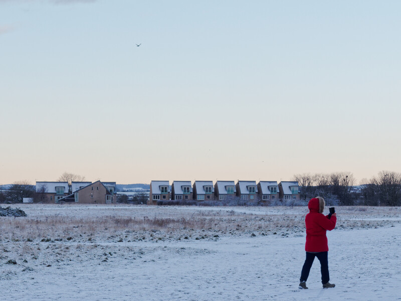 Person in red jacket standing alone on snowy beach with row of beachfront buildings in distance.