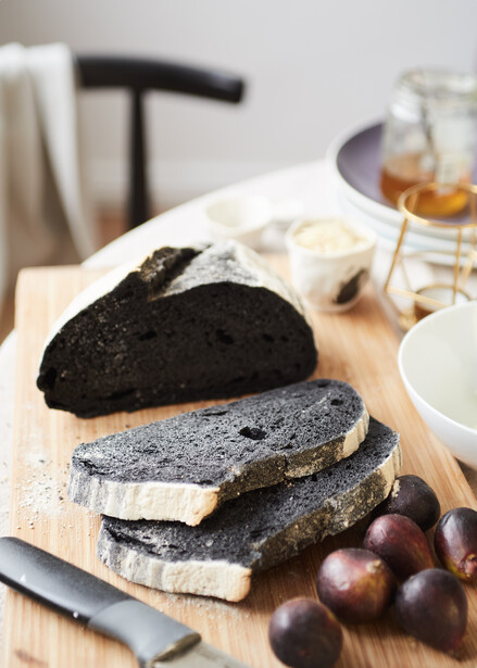 Sliced charcoal bread arranged on parchment paper with butter and grapes on wooden surface.