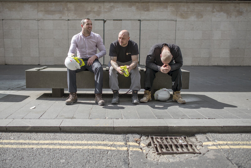 Three men sitting on a concrete bench holding yellow flowers against a gray wall.