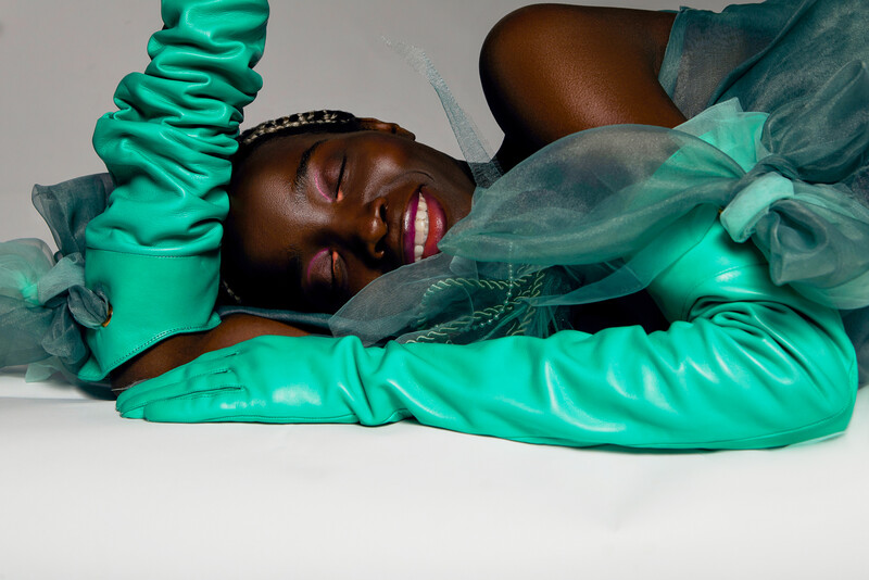 Person lying down wearing bright green latex fabric and sheer teal overlay, photographed from above against a white background.