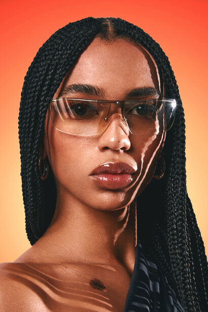 Portrait of a woman wearing clear-framed glasses and earbuds against a warm orange gradient background.