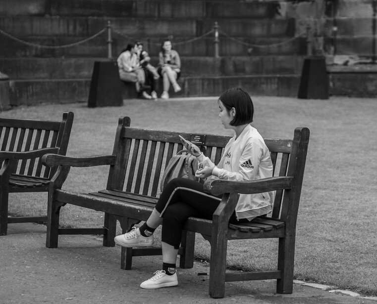 Woman sitting on a bench reading a newspaper in a public courtyard, with other figures visible in soft-focused background.