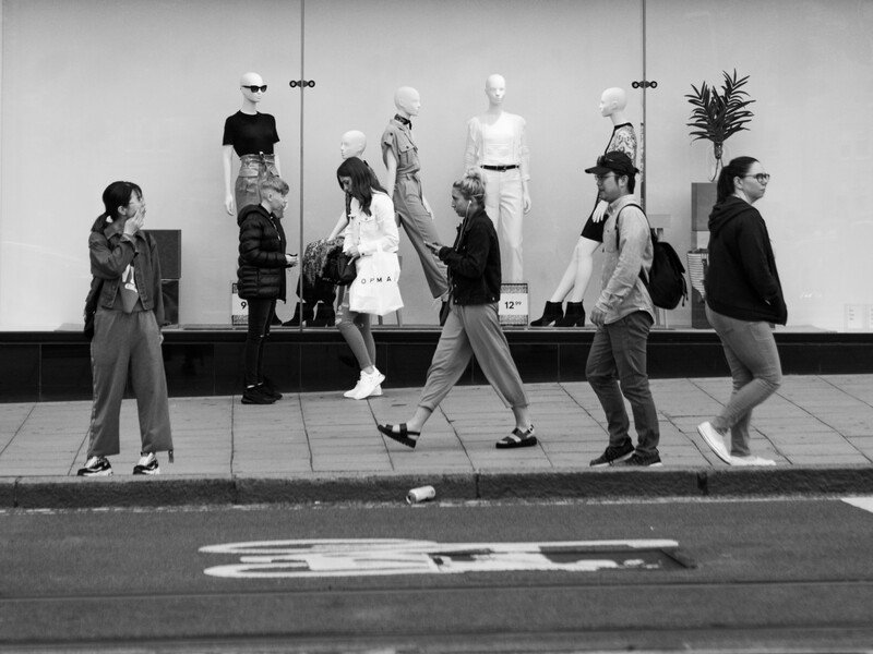 Street-level black and white photograph of pedestrians walking past a storefront window displaying mannequins in various outfits.