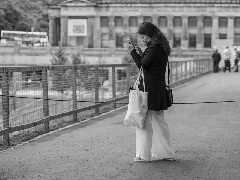 Woman in black coat photographing from a bridge railing overlooking urban architecture.