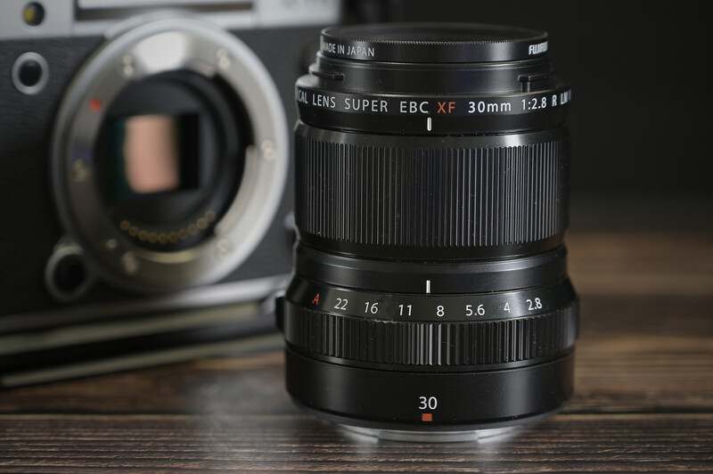 Fujifilm 30mm f/2.8 macro lens with aperture ring and focus scale visible, positioned in front of a camera body.