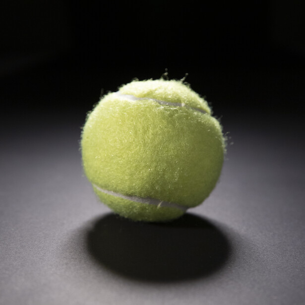 Yellow-green tennis ball with white curved seams on dark surface with shadow.