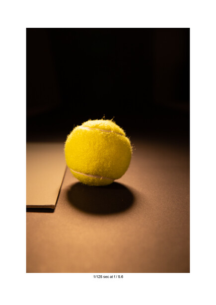 Yellow tennis ball with dramatic shadow on warm tan surface against dark background.