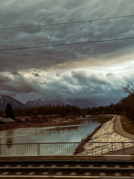 Dramatic storm clouds gather over a river valley with evergreen forests and distant mountains under moody, overcast light.