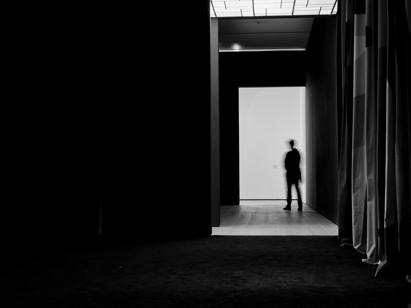 Silhouetted figure standing in a bright white doorway within a dark, minimalist architectural space.