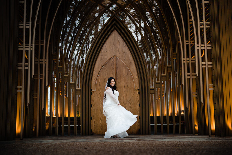 Woman in flowing white dress poses in Gothic cathedral archway with dramatic uplighting.