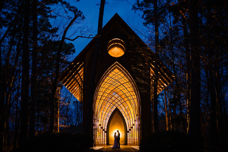 Silhouetted figure standing in the Gothic archway of an illuminated chapel at dusk, surrounded by forest.