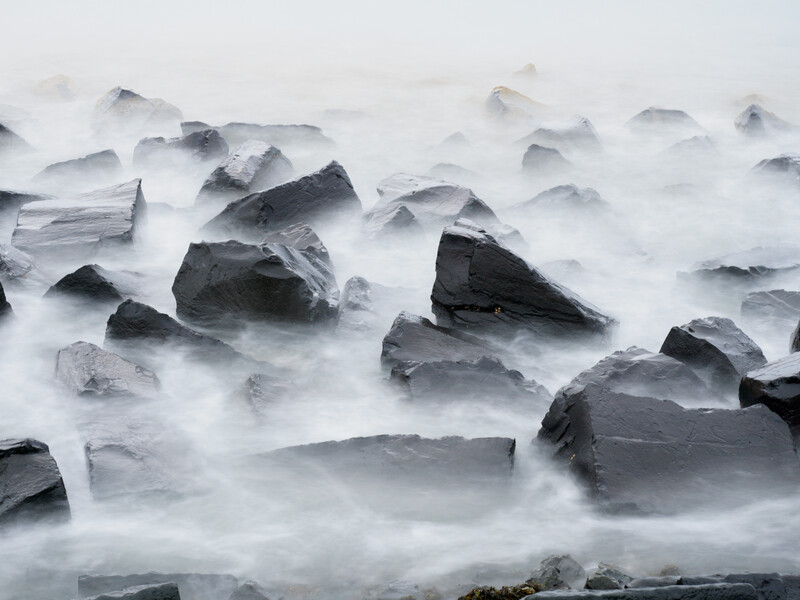 Turbulent ocean waves crashing over dark rocks with silky, motion-blurred white water.