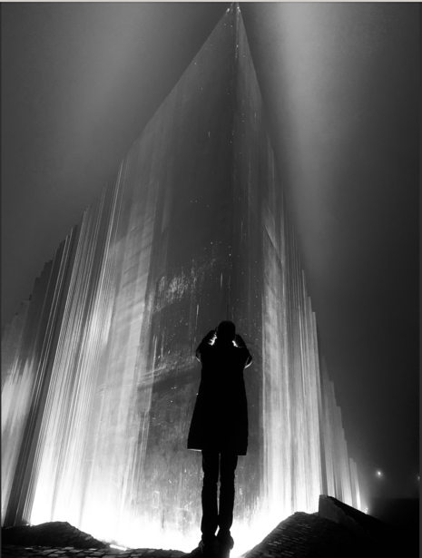Silhouetted figure standing before a massive illuminated architectural structure shot from below.