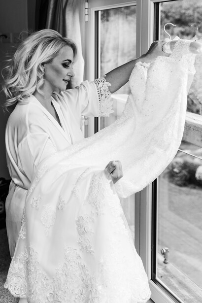 Bride in lace wedding dress examining sheer veil by window in black and white portrait.