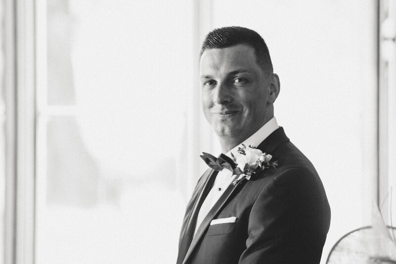 Groom in black tuxedo with bow tie and boutonniere smiling at camera by window.