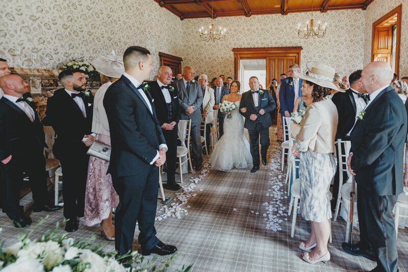 Wedding ceremony in an ornate room with guests seated on either side of an aisle as the bride and groom stand at the altar.