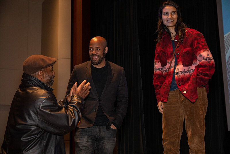 Three people conversing on a stage with warm theatrical lighting and dark curtains behind them.