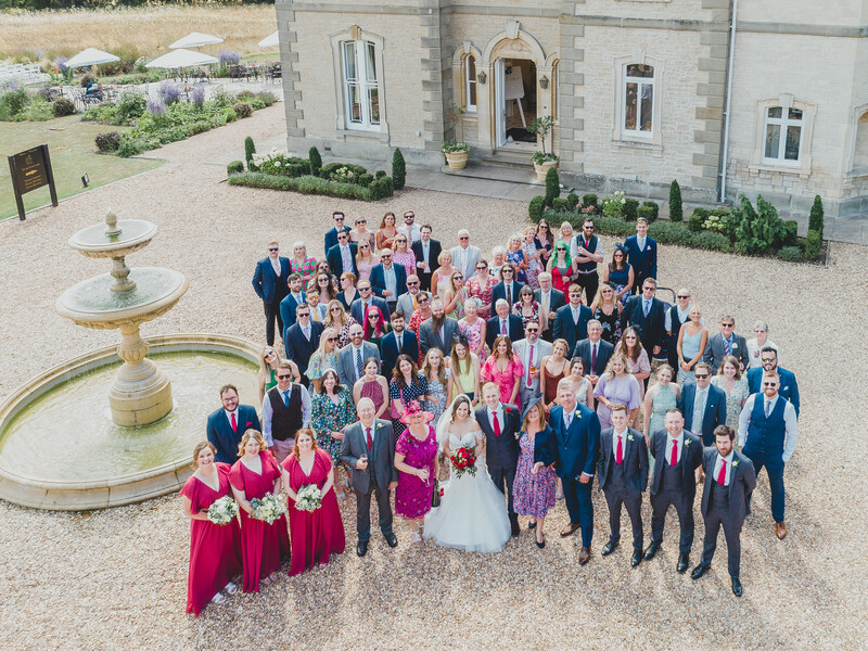 Large wedding party of approximately 80 guests arranged in rows on a gravel courtyard in front of a stone building.