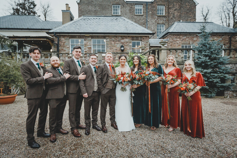 Wedding party of eleven people in formal attire standing in front of a stone country house.
