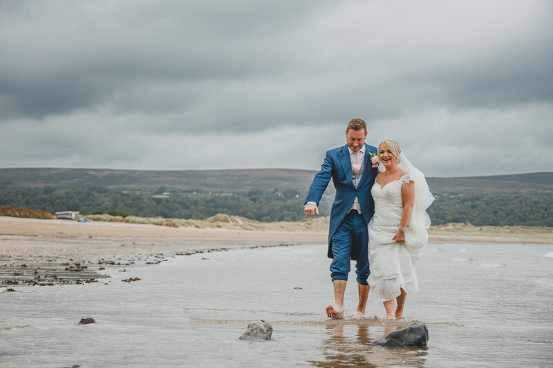 Couple in formal attire wading through shallow water on a beach at sunset.