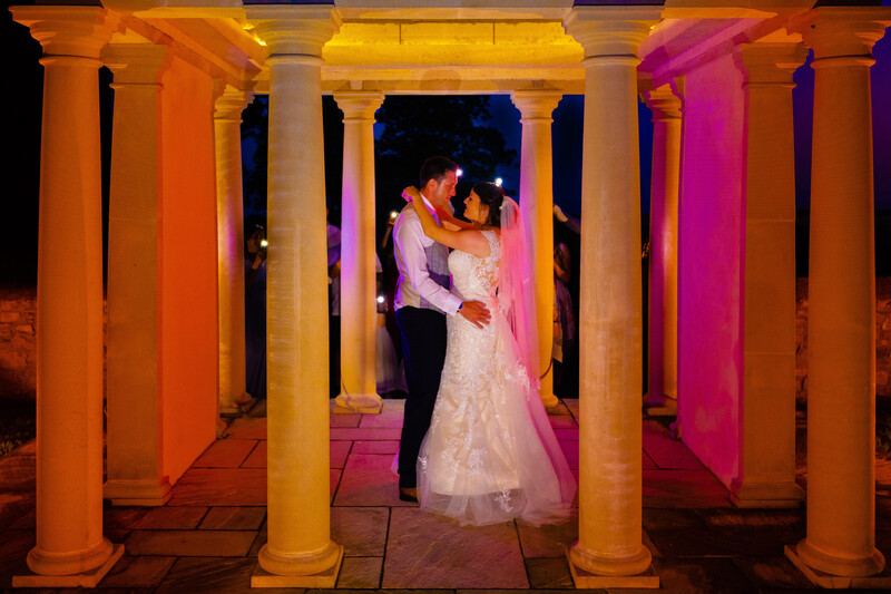 Bride and groom embracing under illuminated columns at night.