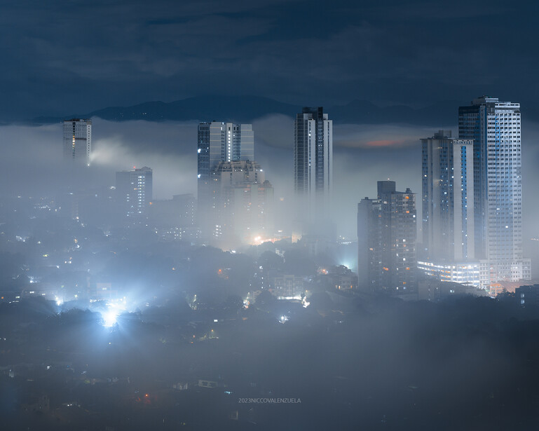 City skyline shrouded in dense fog and mist during twilight, with illuminated skyscrapers emerging from the haze.