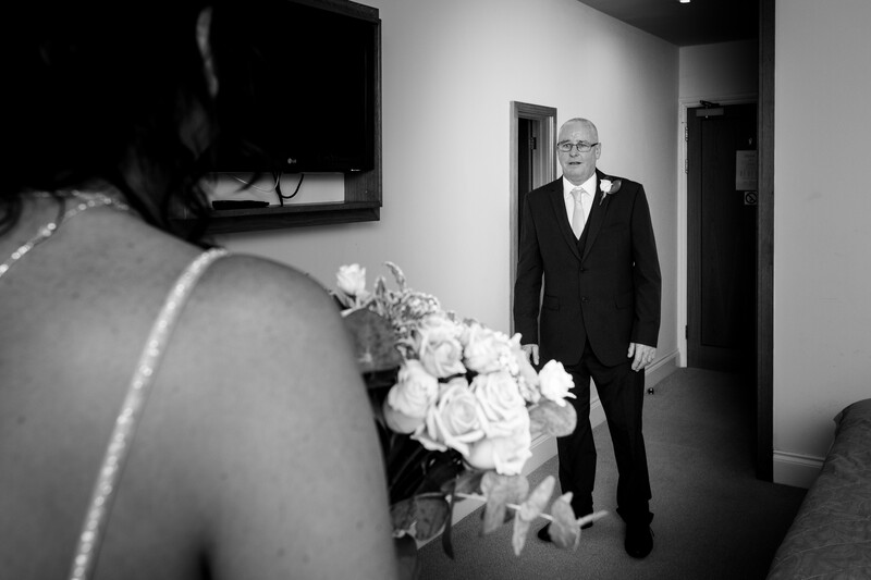 Groom in dark suit watches bride approach down hallway, with white floral bouquet visible in foreground.