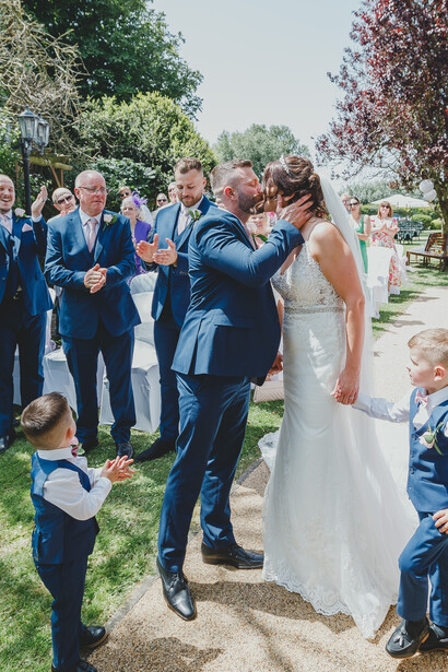 Groom laughing with bride during outdoor garden wedding ceremony surrounded by guests.