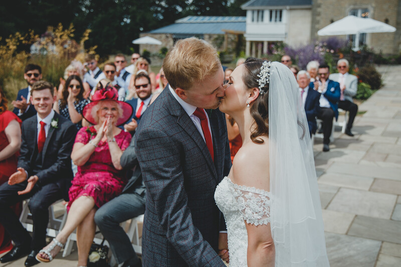 Groom and bride kiss during outdoor wedding ceremony with seated guests visible in background.