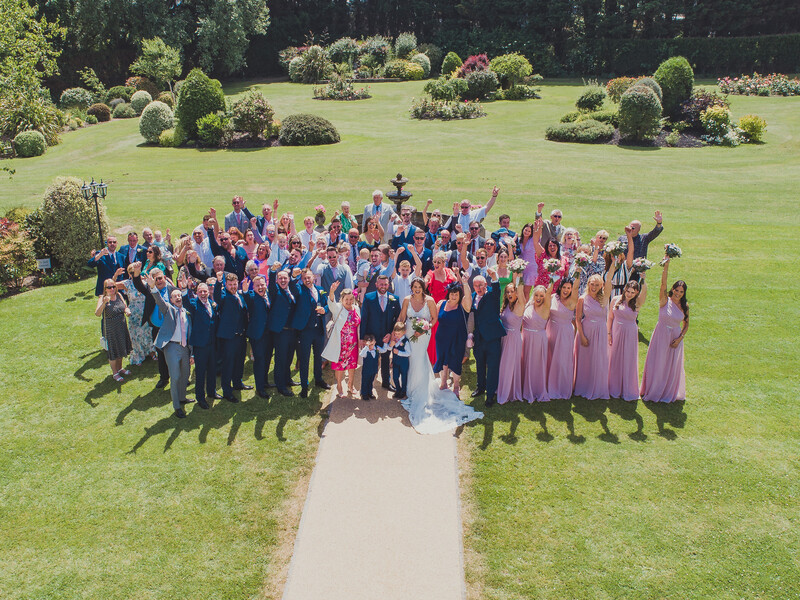 Large wedding party posed in formal attire on manicured lawn during daytime.