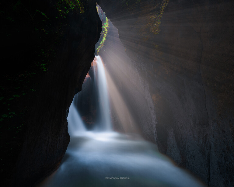 Narrow slot canyon with a flowing stream at its base, illuminated by sunlight streaming through the opening above.