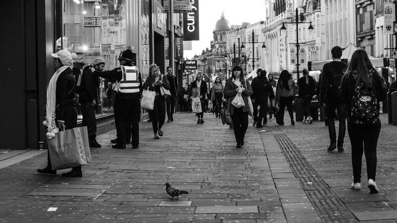Busy urban shopping street captured in black and white, with pedestrians and a lone pigeon on the pavement.