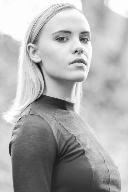 Woman with long blonde hair wearing dark fitted top, looking over shoulder in contemplative pose.