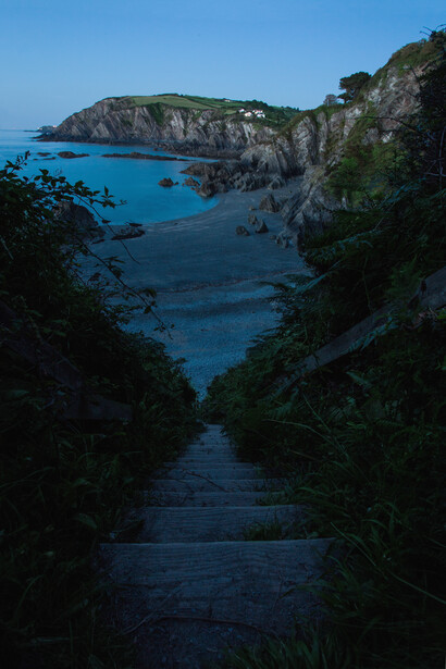 Narrow stone stairway framed by lush vegetation descends toward a rocky cove with turquoise water and dramatic cliffsides.