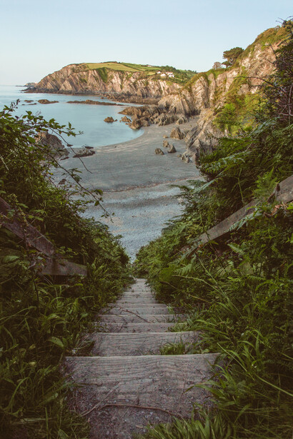 Wooden boardwalk framed by lush vegetation leading toward a rocky coastal cove with turquoise water.