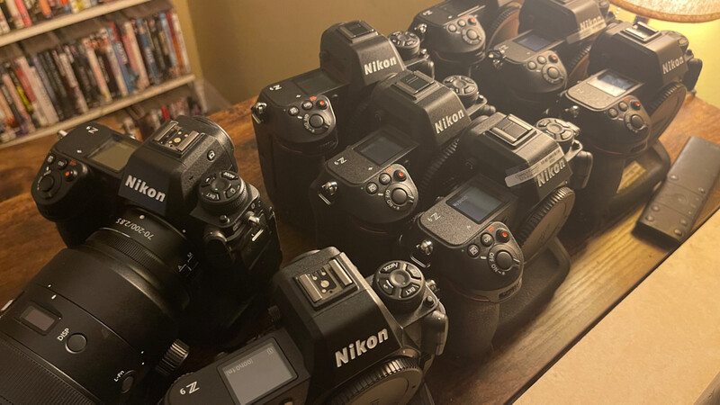Collection of Nikon DSLR cameras and lenses arranged on a wooden table with bookshelves in the background.