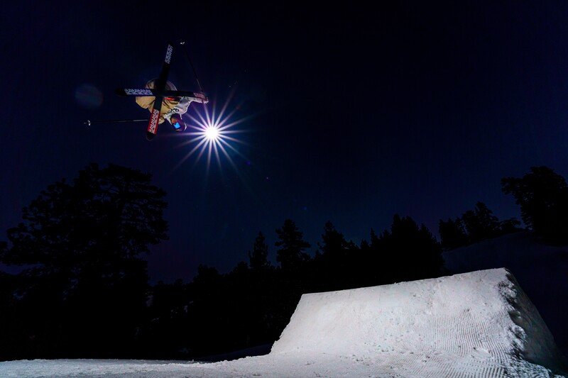 Skier performing aerial trick against night sky with bright moon and evergreen trees below snowy slope.