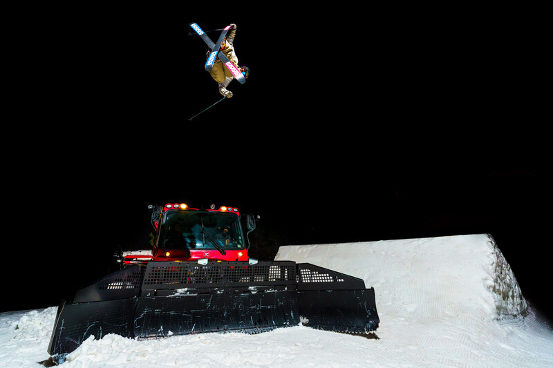 Snowboarder launching into air above a snow-covered grooming vehicle at night.