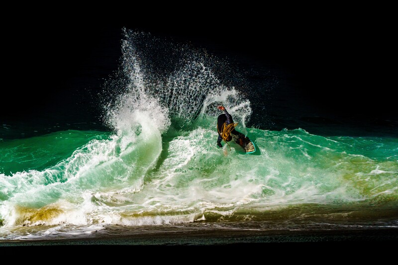 Surfer riding a large wave with dramatic water spray against a black night sky.