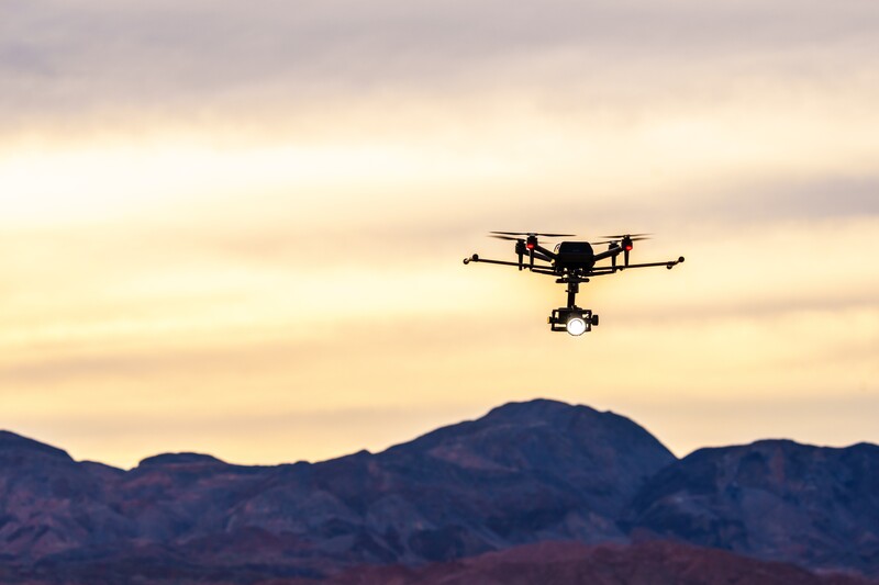 Hexacopter drone silhouetted against golden sunset sky above desert mountains.