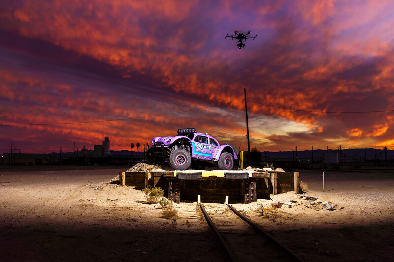Off-road racing truck mounted on concrete platform under dramatic orange and purple sunset sky with drone flying overhead.