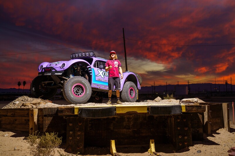Person in pink shirt standing on modified truck against dramatic sunset sky with orange clouds.