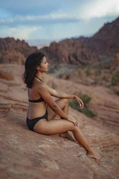 Woman in bikini sitting on sandy terrain with layered mountains in the background.