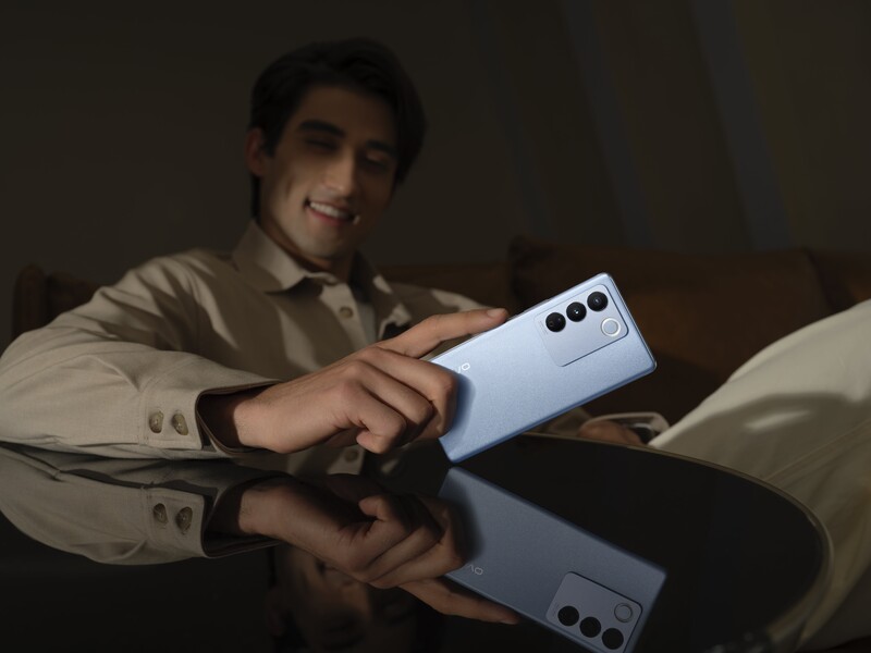 Man holding a light blue smartphone with triple camera setup, reflected on a dark surface.