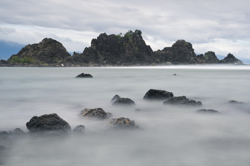 Rocky islands shrouded in mist rising from calm ocean waters under cloudy sky.