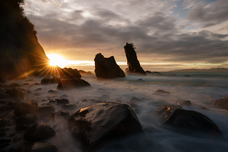 Rocky sea stacks silhouetted against a golden sunrise over turbulent ocean waters.