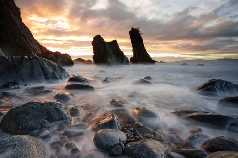 Dramatic seascape with towering rock formations silhouetted against a golden sunset sky, surrounded by swirling mist and boulders.