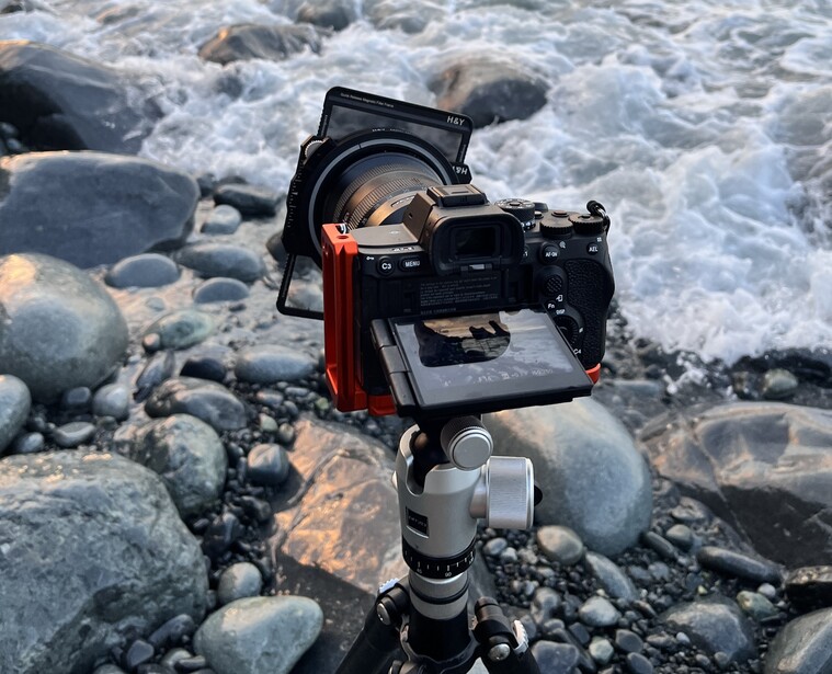 Mirrorless camera on tripod mounted at rocky shoreline with waves crashing in background.
