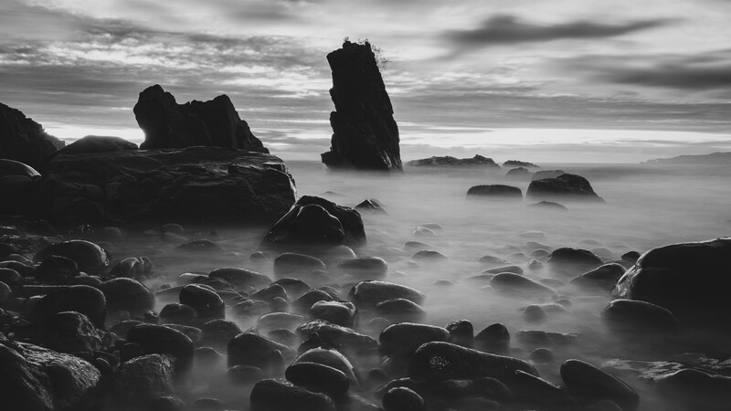 Rocky coastline with tall sea stacks and boulders shrouded in misty surf beneath dramatic clouds.
