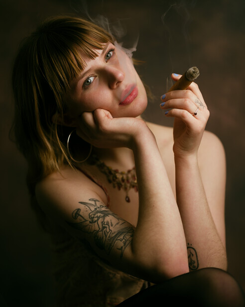 Woman with bangs and shoulder tattoos holding a small wooden figurine, gazing upward thoughtfully.